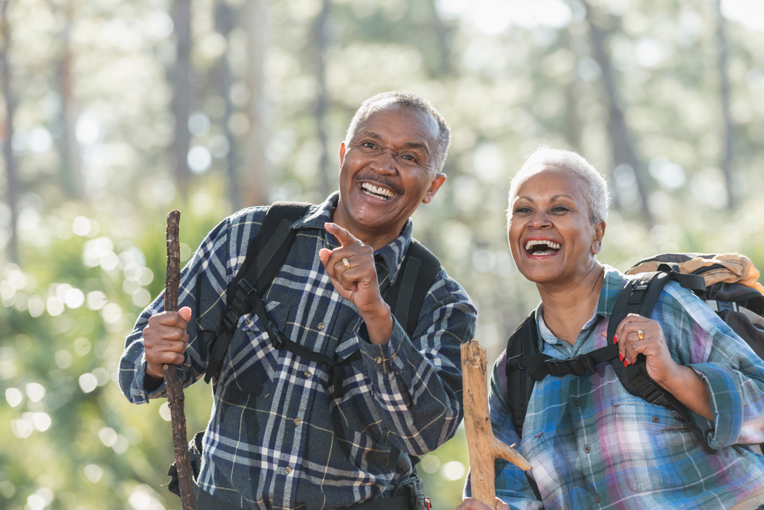 An African American senior couple hiking through the woods carrying backpacks and walking sticks. They are smiling and watching something the man is pointing at.